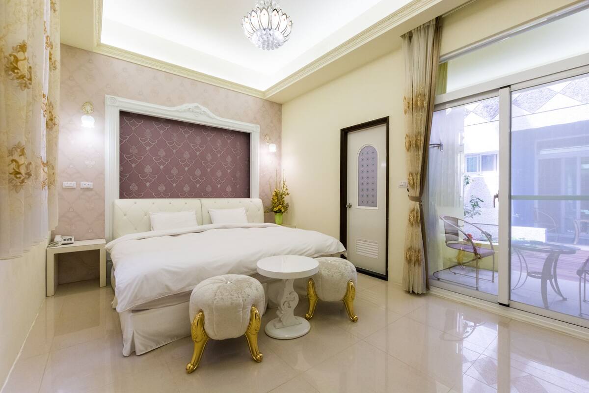 Quarto com uma cama grande, uma mesinha redonda, dois pufes, um candelabro e uma porta de vidro deslizante para área externa.