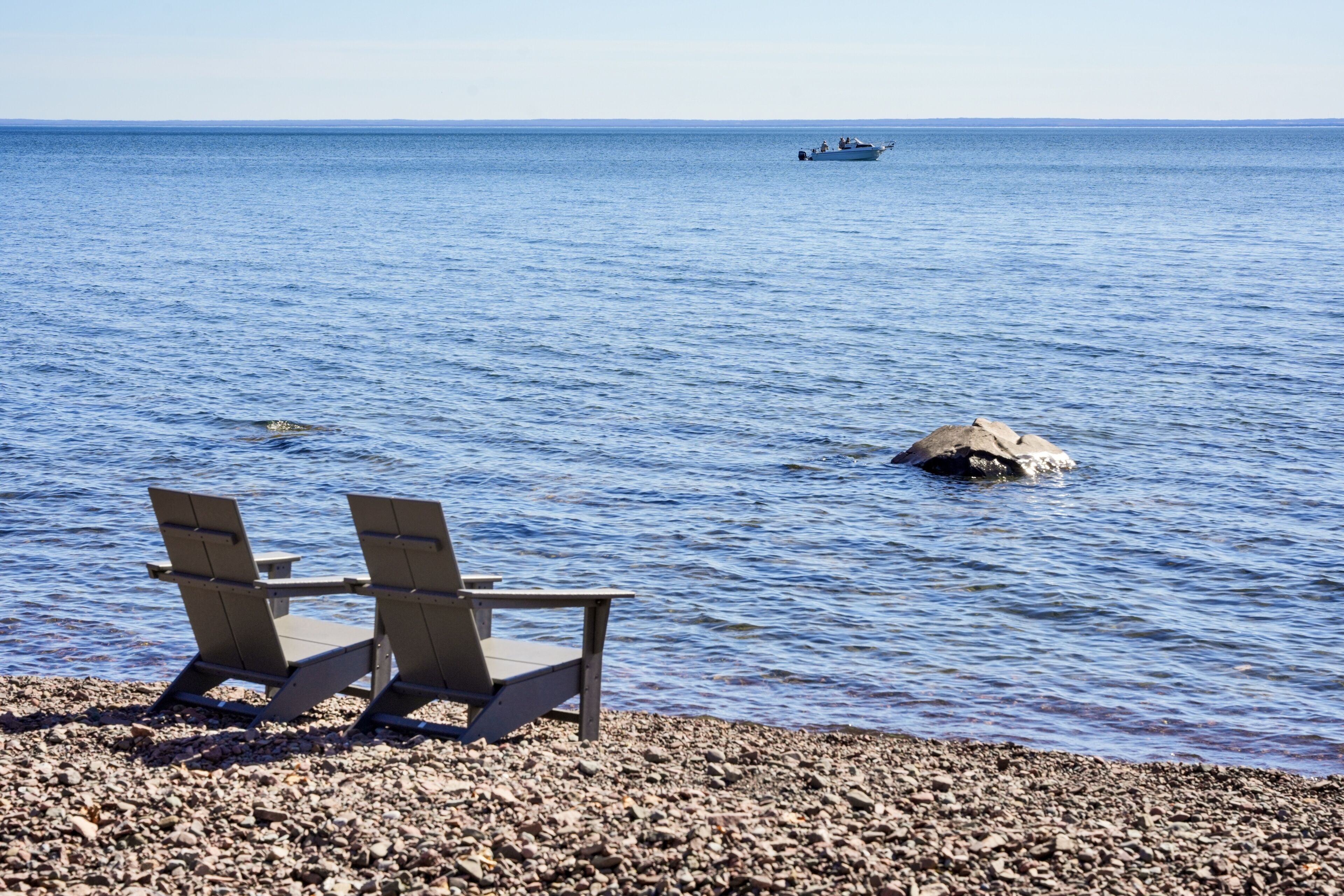 A beach with two wooden chairs facing the sea, a rocky shore, and a boat in the distance.