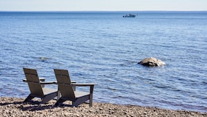 A beach with two wooden chairs facing the sea, a rocky shore, and a boat in the distance. - Sisu Shores: Lakefront Home with Private Beach and Wood Burning Sauna (Duluth)