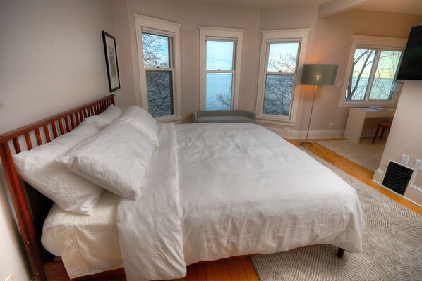 A bedroom with a large bed, multiple windows, and a view of the water. - Sisu Shores: Lakefront Home with Private Beach and Wood Burning Sauna (Duluth)