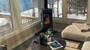 A cozy living room with a wood-burning stove, large windows with a view of a snowy landscape, and a comfortable seating area. - Sisu Shores: Lakefront Home with Private Beach and Wood Burning Sauna (Duluth)
