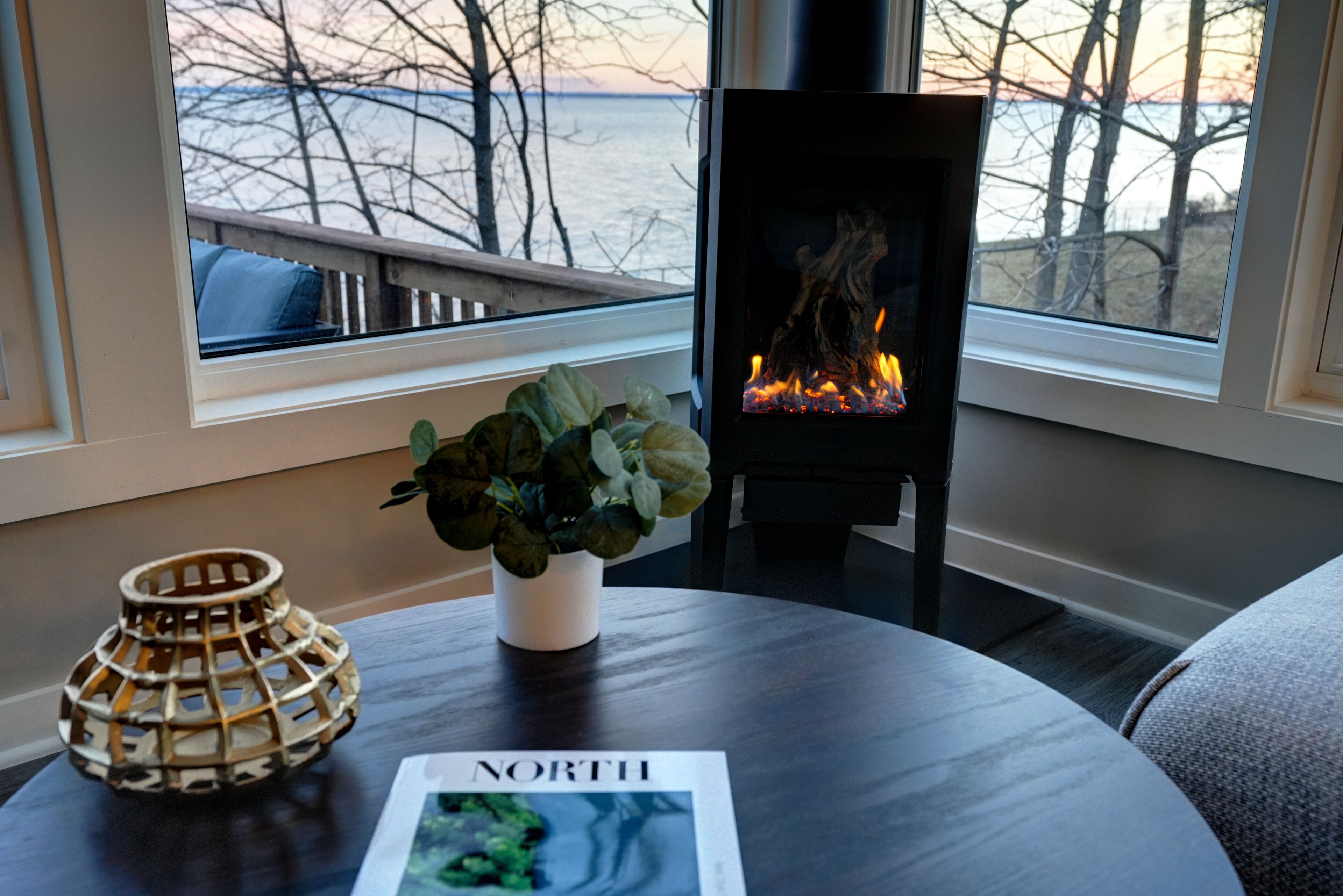 A cozy room with a fireplace, a window with a view of trees and water, a round table with a plant and a magazine, and a decorative lamp.