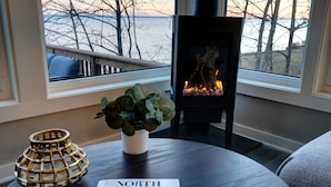 A cozy room with a fireplace, a window with a view of trees and water, a round table with a plant and a magazine, and a decorative lamp. - Sisu Shores: Lakefront Home with Private Beach and Wood Burning Sauna (Duluth)