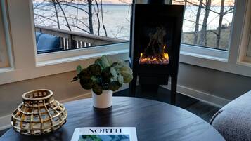 A cozy room with a fireplace, a window with a view of trees and water, a round table with a plant and a magazine, and a decorative lamp.
