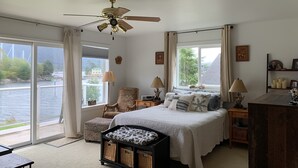 A bedroom with a bed, two lamps, a chair, a desk, and a view of a body of water through large windows.