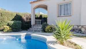 Ein Schwimmbecken mit geometrischem Fliesenmuster, eine Terrasse mit Gartenmöbeln sowie ein Haus mit Naturstein-Fassade und einem Fenster mit weißen Sprossen.