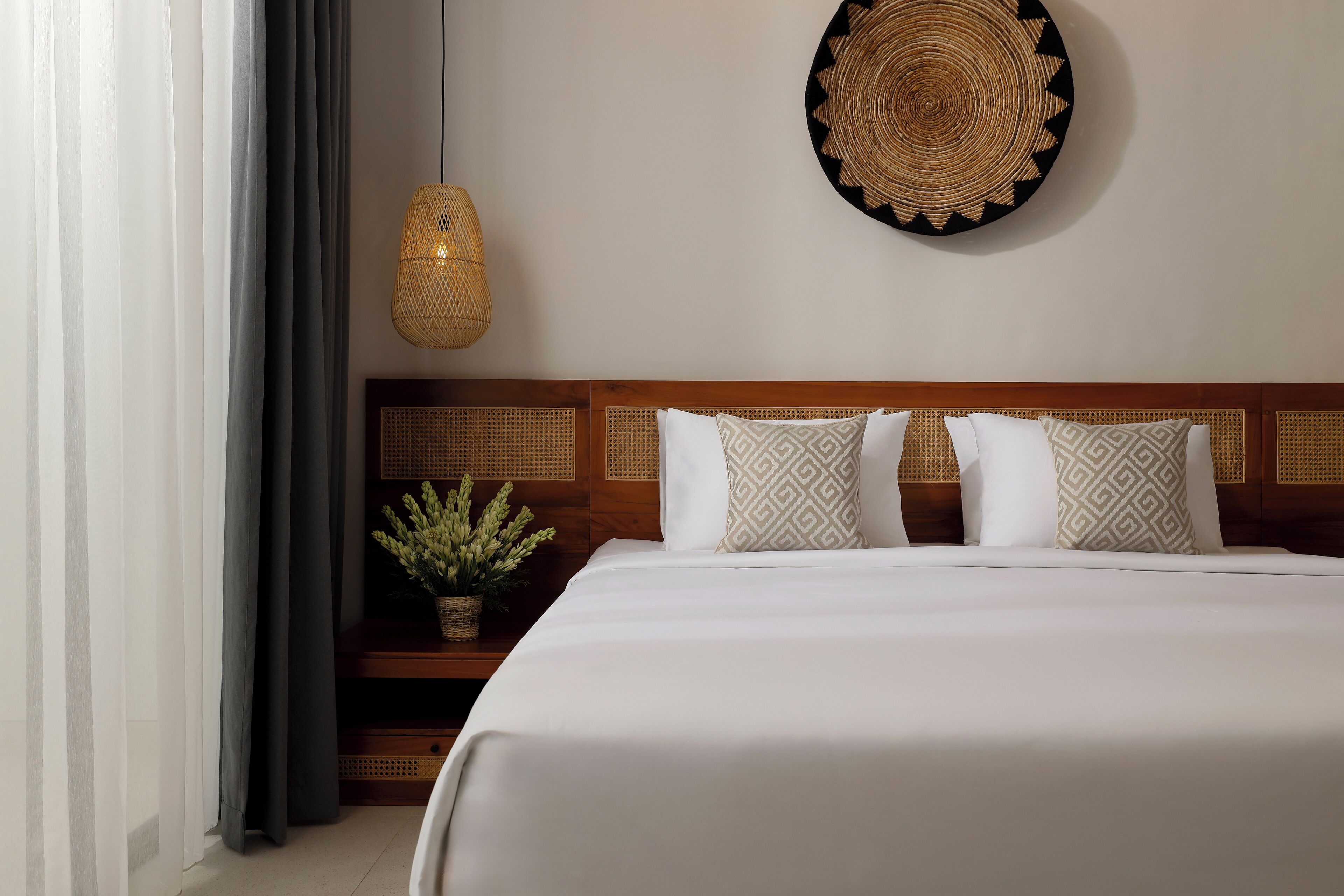 A neatly made bed with a wooden headboard, a woven wall hanging, and a hanging light fixture.