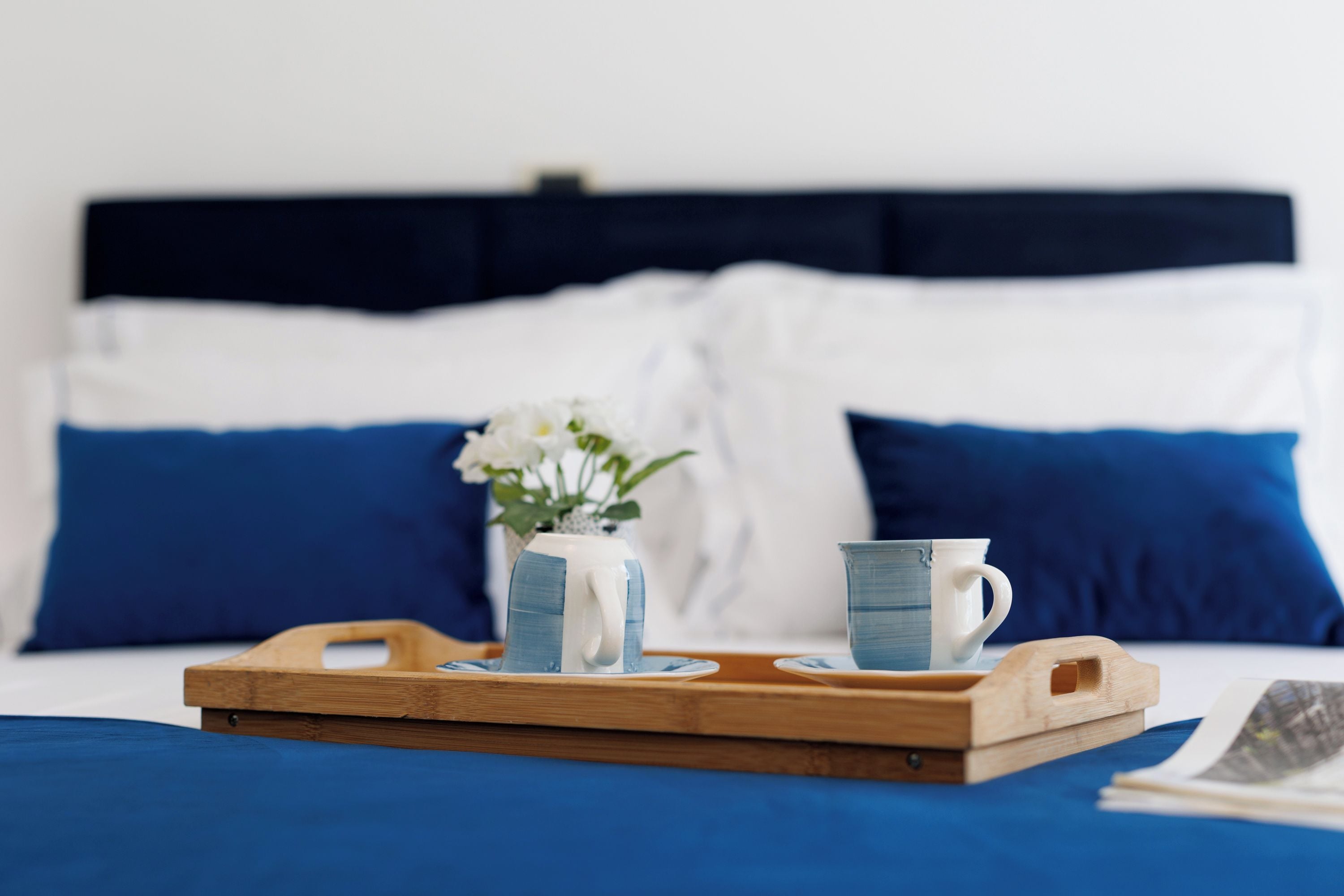 A bed with blue bedding, a wooden tray with two ceramic mugs, and a vase with flowers.