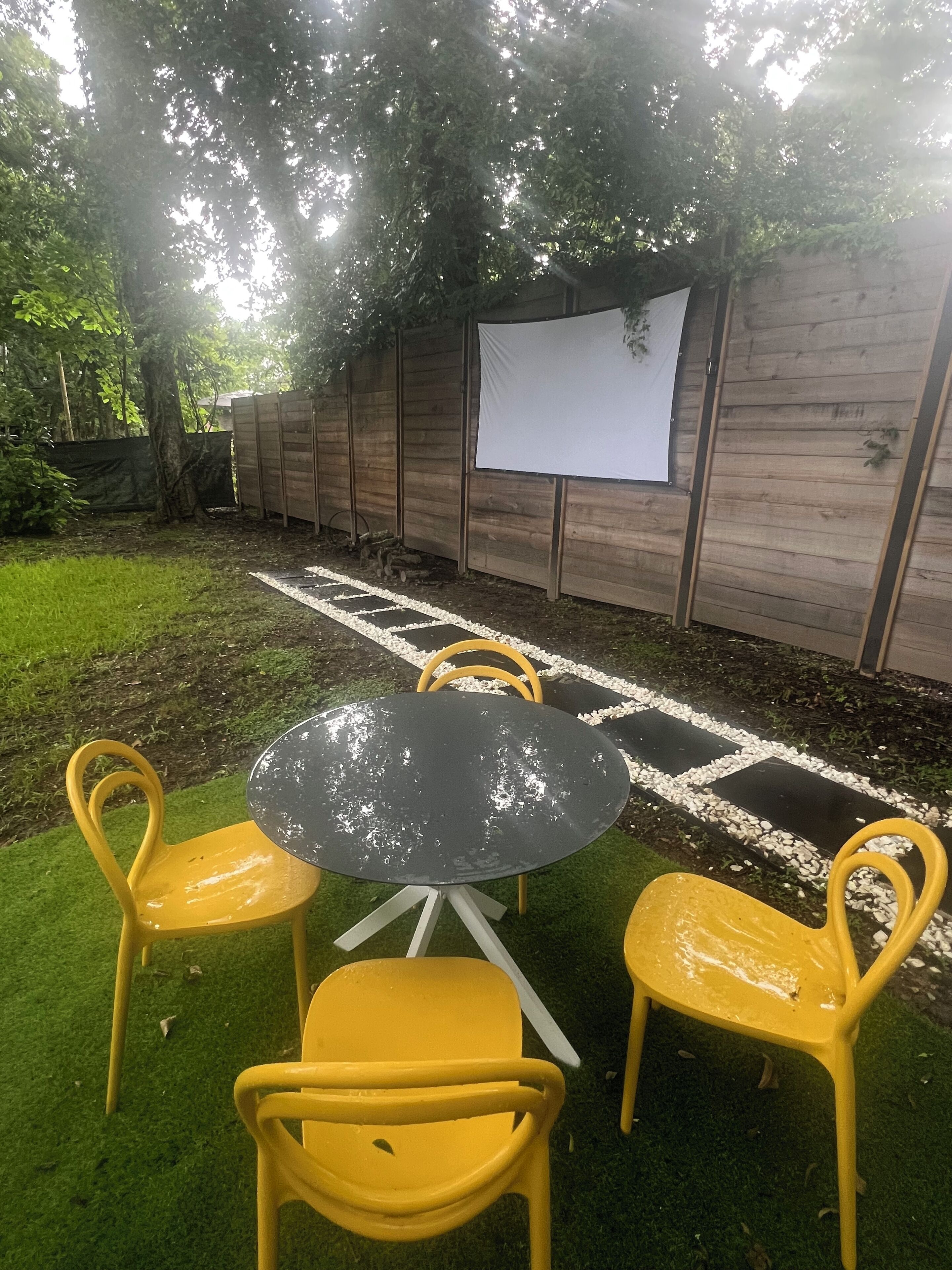 Un espace extérieur aménagé avec une table ronde et des chaises jaunes, entouré d’une clôture en bois et d’un écran blanc.