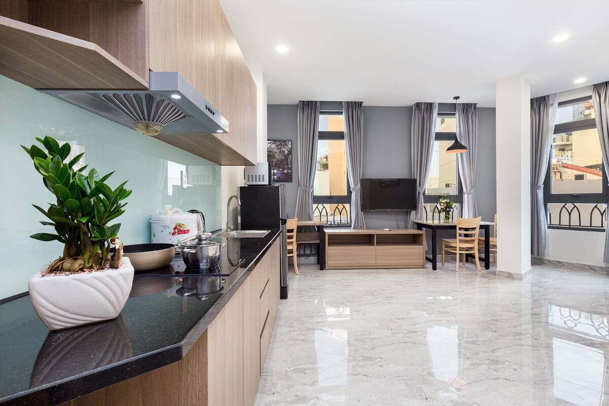A modern kitchen with a black countertop, wooden cabinets, and a built-in stove. There is a dining area with a table and chairs, and a large window with curtains.