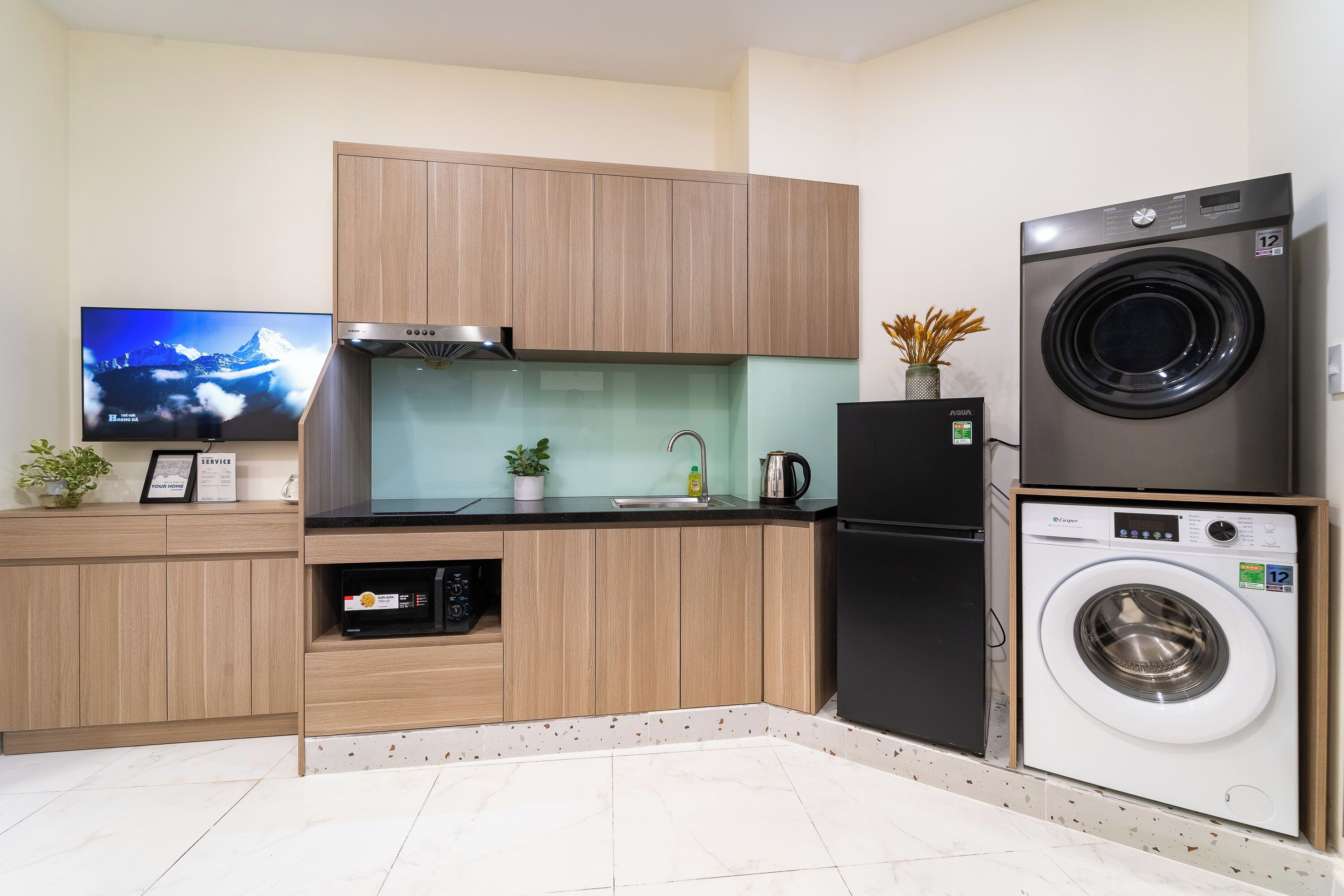 A compact kitchen with wooden cabinets, a built-in washing machine, a refrigerator, and a flat-screen TV.