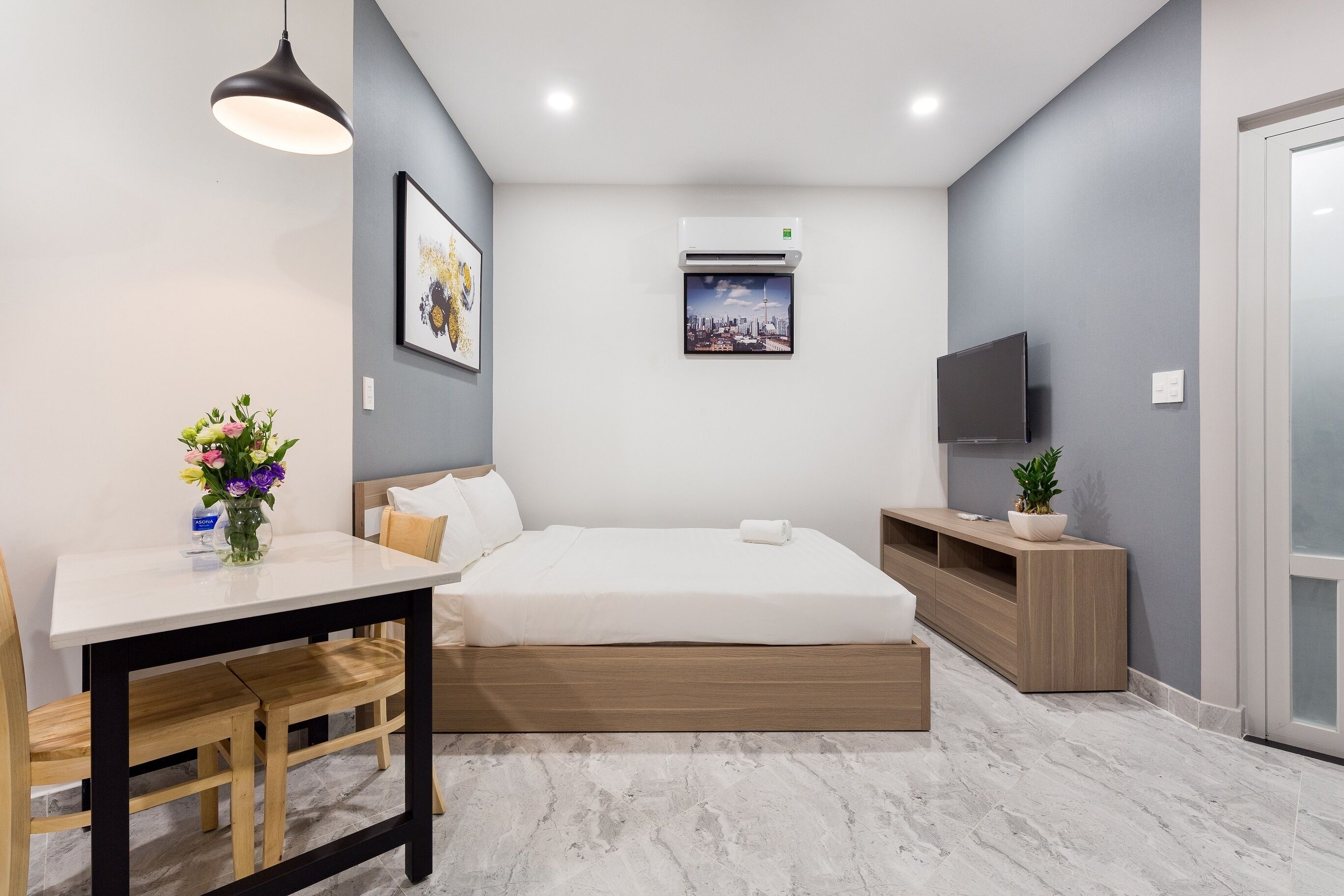 A modern hotel room with a bed, a desk with chairs, a TV, and a wall-mounted air conditioner.