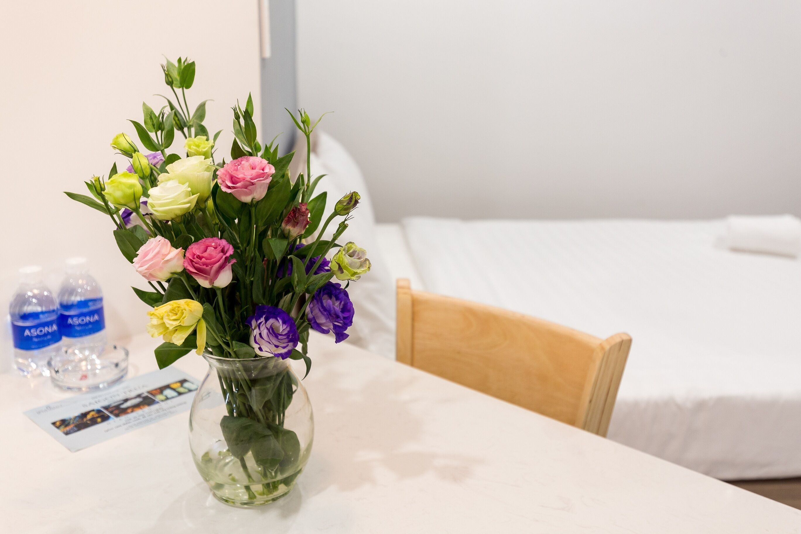 A hotel room with a bed, a wooden chair, a table with a vase of flowers, and two bottles of water.