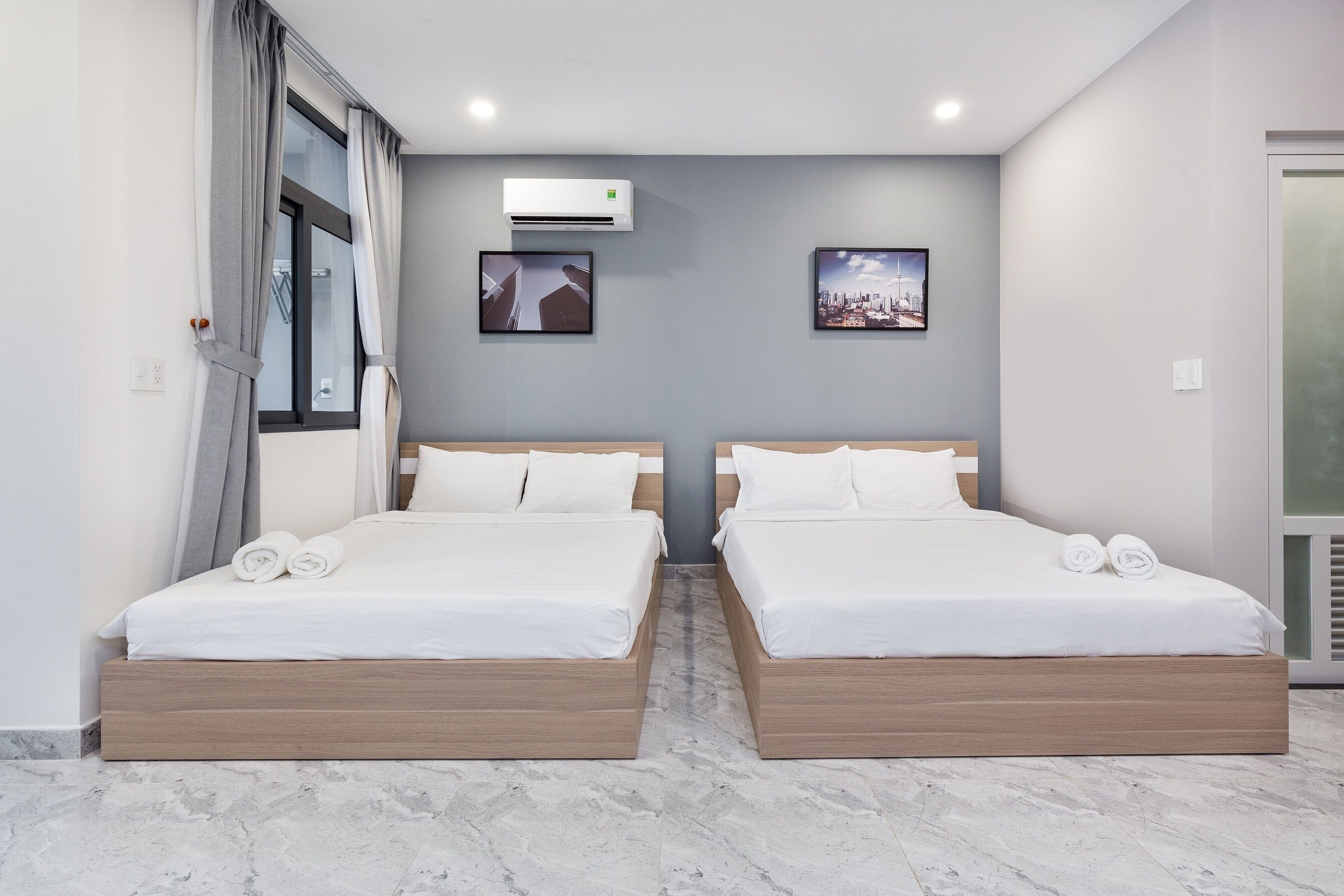 Una habitación de hotel moderna con dos camas, un ventanal con cortinas, un equipo de aire acondicionado y dos cuadros enmarcados en la pared.