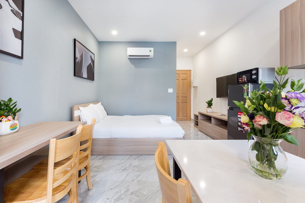 A modern hotel room with a bed, a desk, and a dining area. There is a kitchen area with a refrigerator and a microwave.
