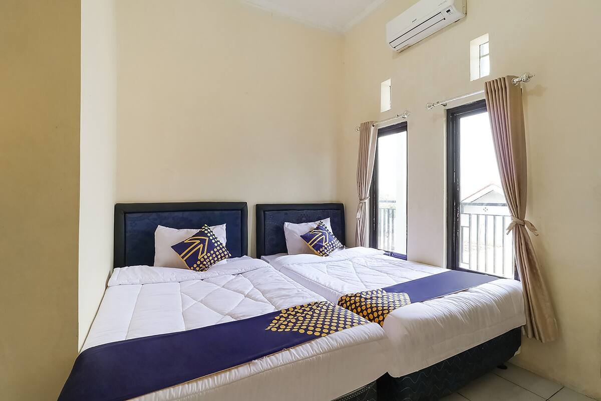 Una cama doble con una colcha azul y blanca, dos almohadas con diseño geométrico y una ventana con cortinas.
