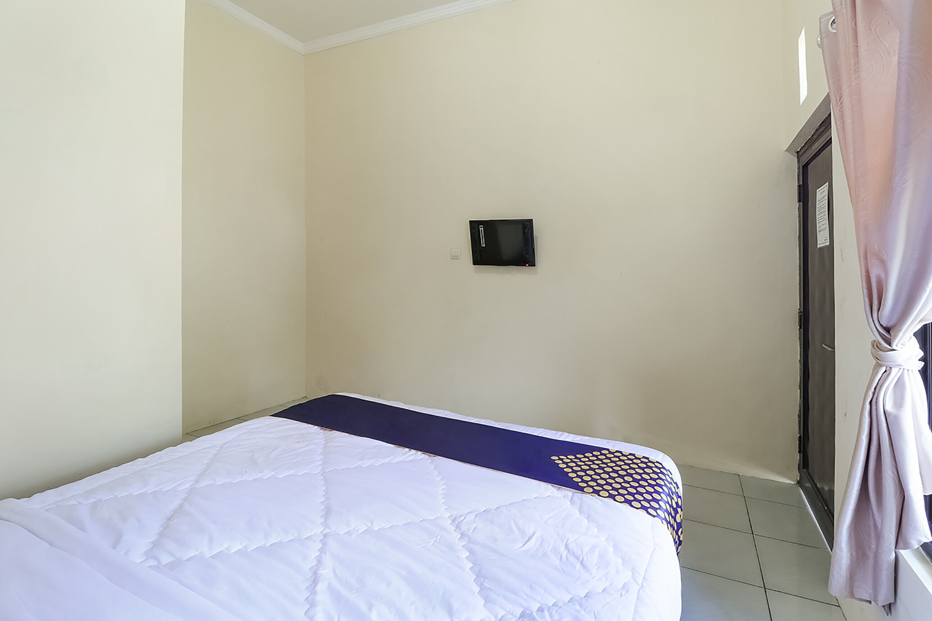 Un lit simple avec une couette blanche et un oreiller à motifs bleus et jaunes, un petit appareil noir fixé au mur et un rideau attaché au cadre de la porte.