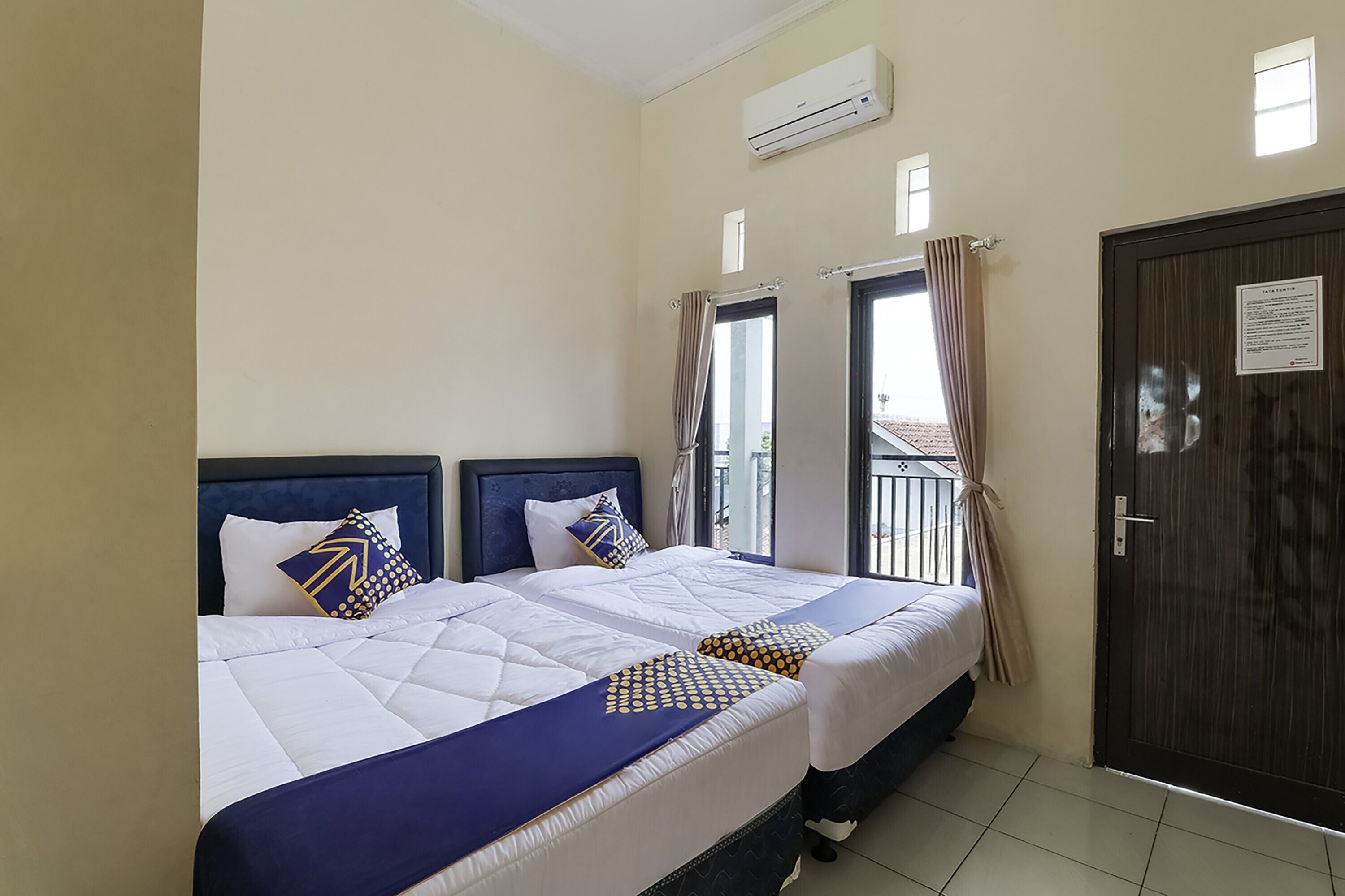A hotel room with two beds, a dark wooden door, and an air conditioning unit on the wall.