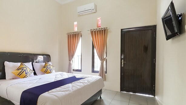 A bedroom with a bed, curtains, a door, and a wall-mounted TV. - SPOT ON 3859 Sweet Candy 2NearResinda park mall (Karawang)