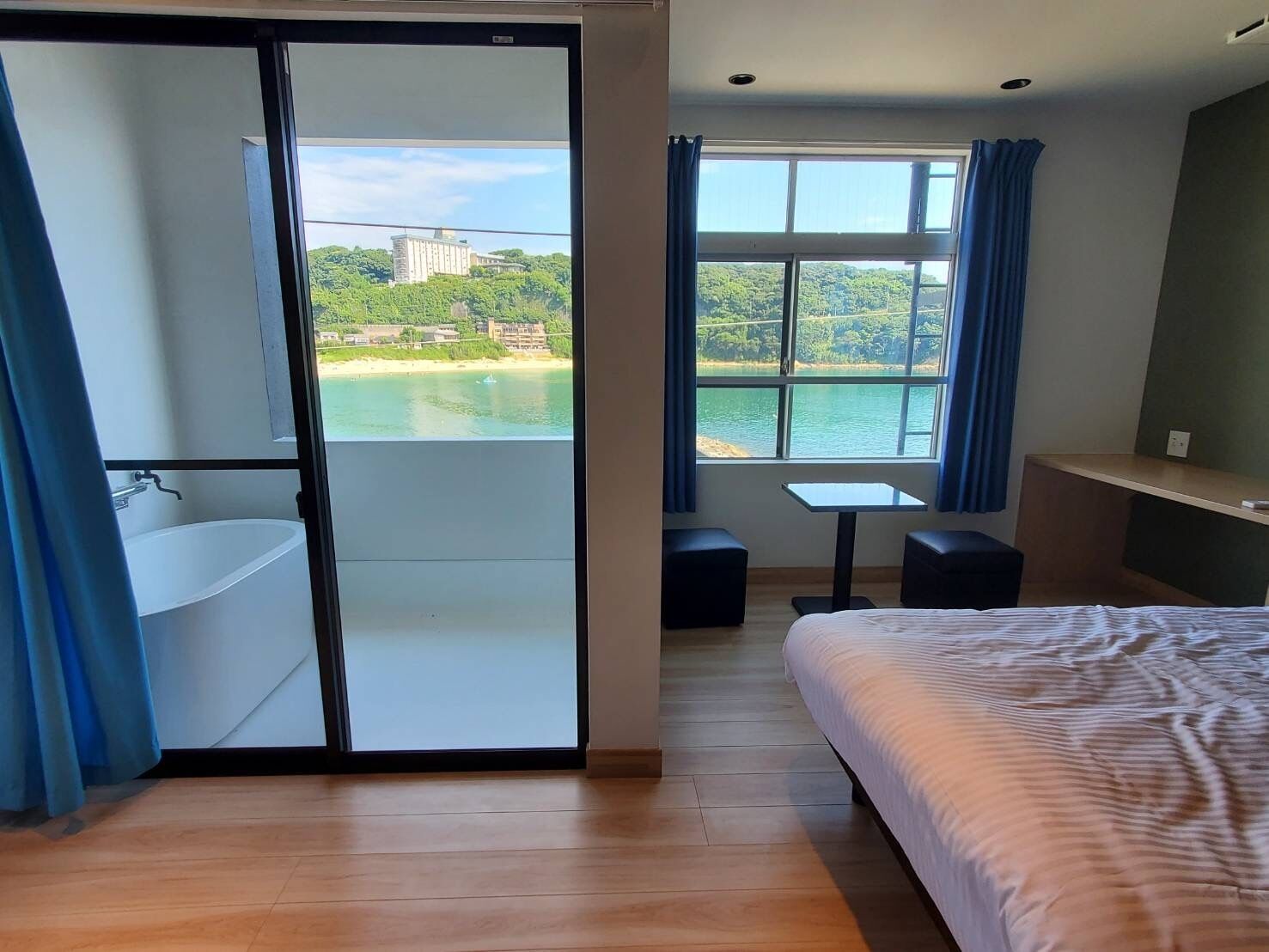 Ein Schlafzimmer mit Bett, kleinem Tisch, Stuhl, Schreibtisch, Fenster mit Blick auf ein Gewässer und Gebäude sowie ein Badezimmer mit Waschbecken und Dusche.