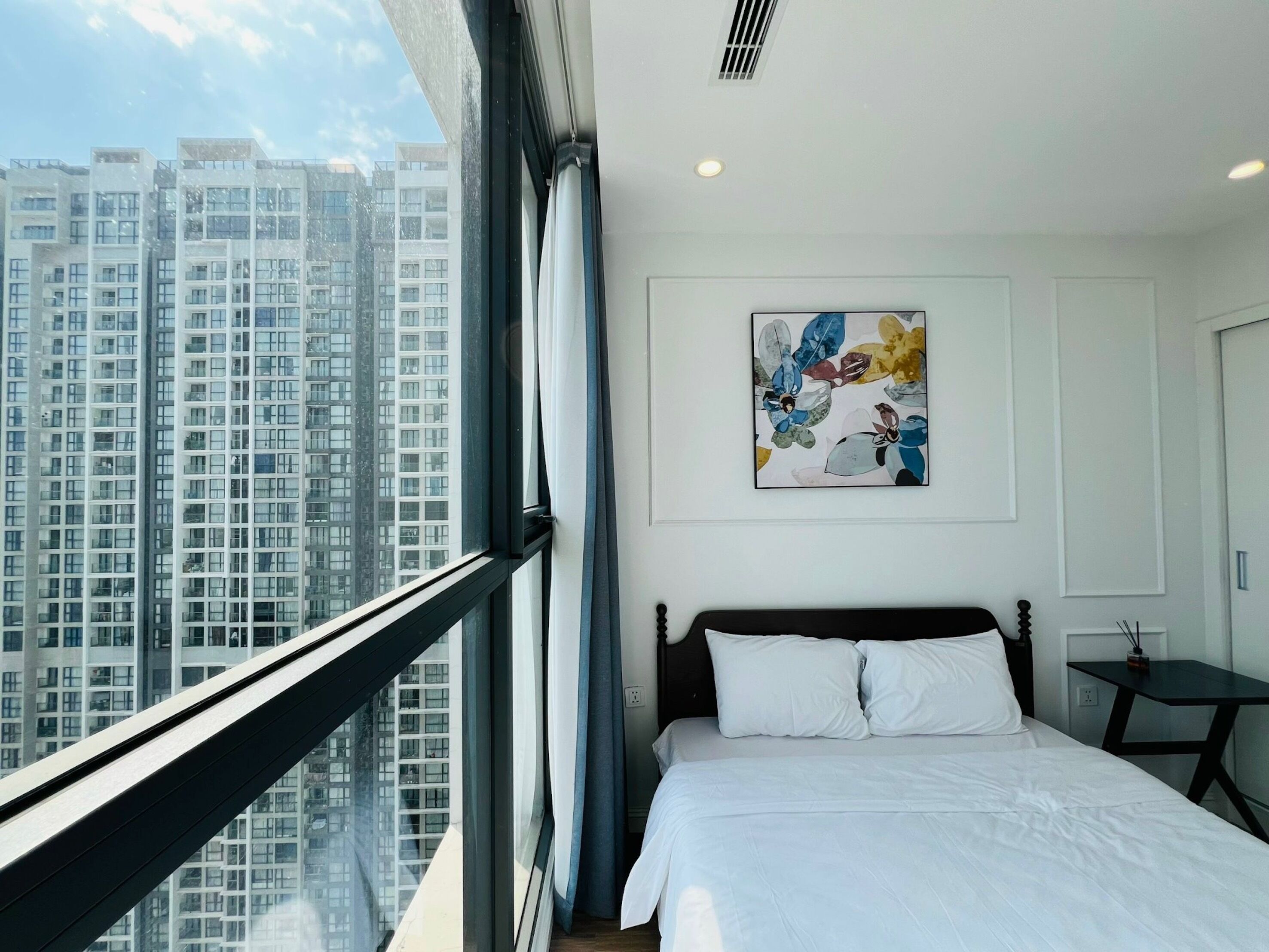 A modern hotel room with a large window, a bed with white linens, a black headboard, and a colorful abstract painting on the wall.