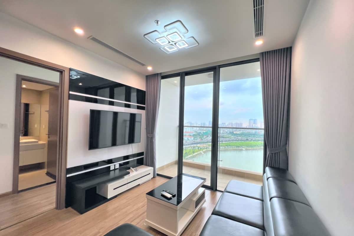 A modern living room with a flat-screen TV, a coffee table, and a sofa. There is a large window with a view of a cityscape.