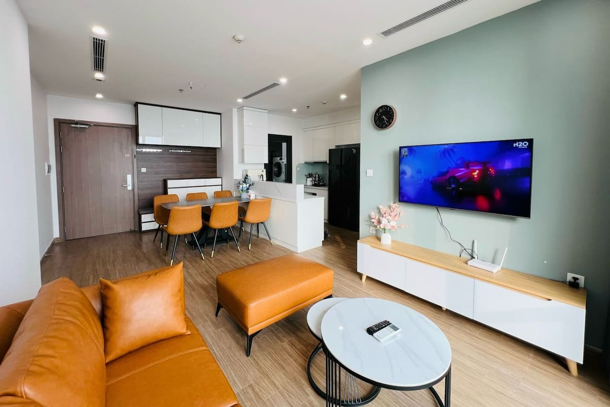 A modern living room with a sofa, ottoman, and a coffee table. There is a dining area with chairs and a table. A TV is mounted on the wall, and a clock is visible.