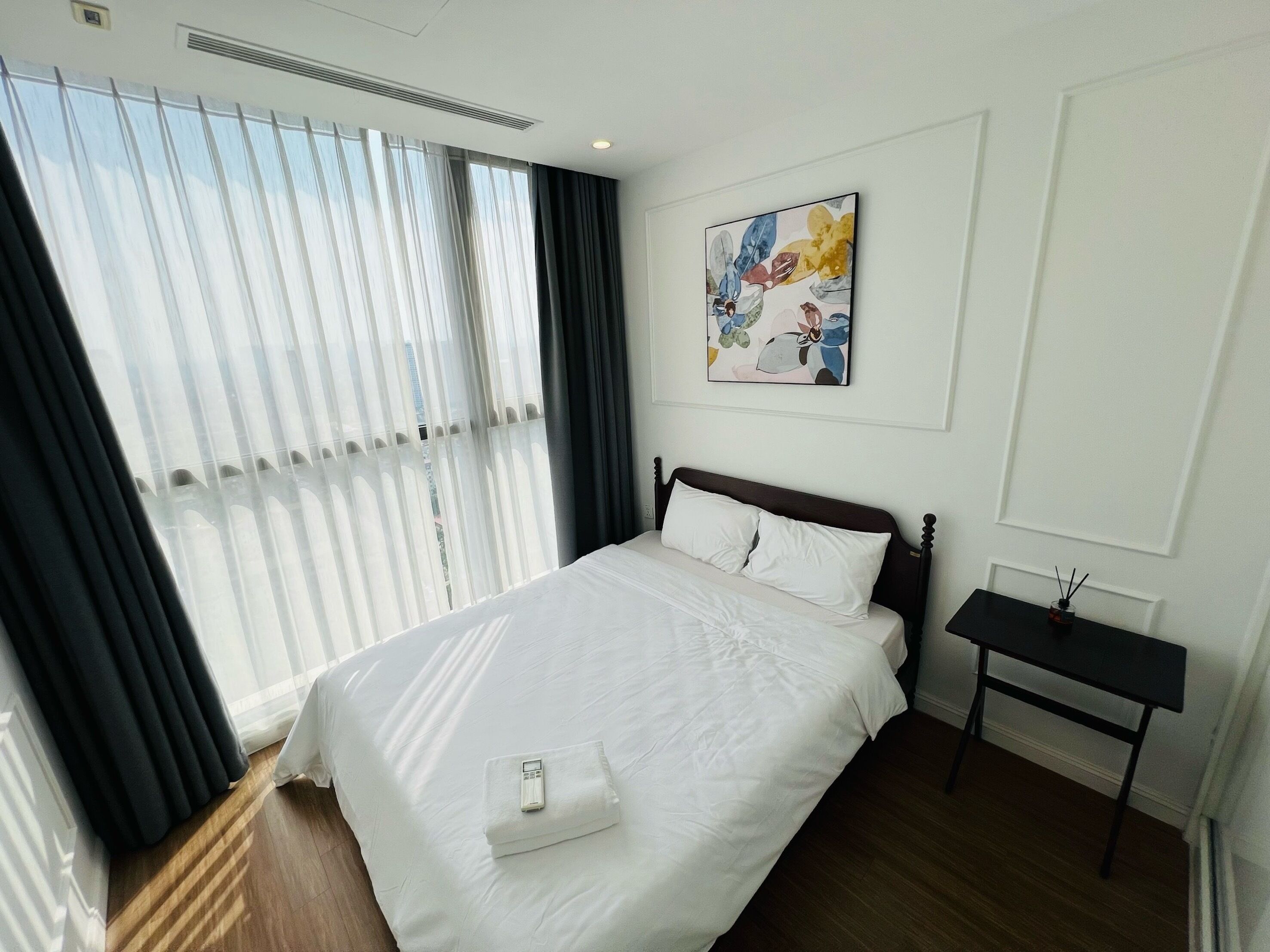 Hotelkamer met een groot bed, nachtkastje, schilderij aan de muur en raam met dunne gordijnen.