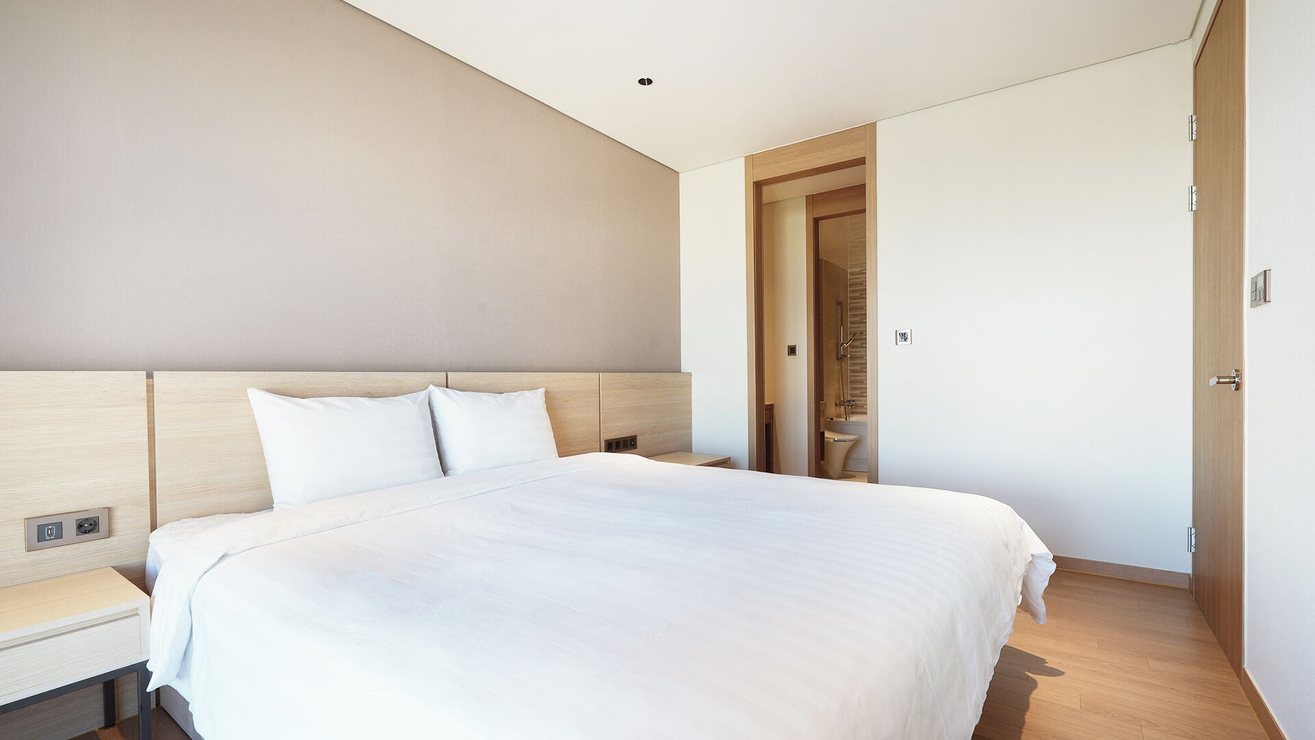 A modern hotel room with a large bed, a bedside table, and a door leading to a bathroom.