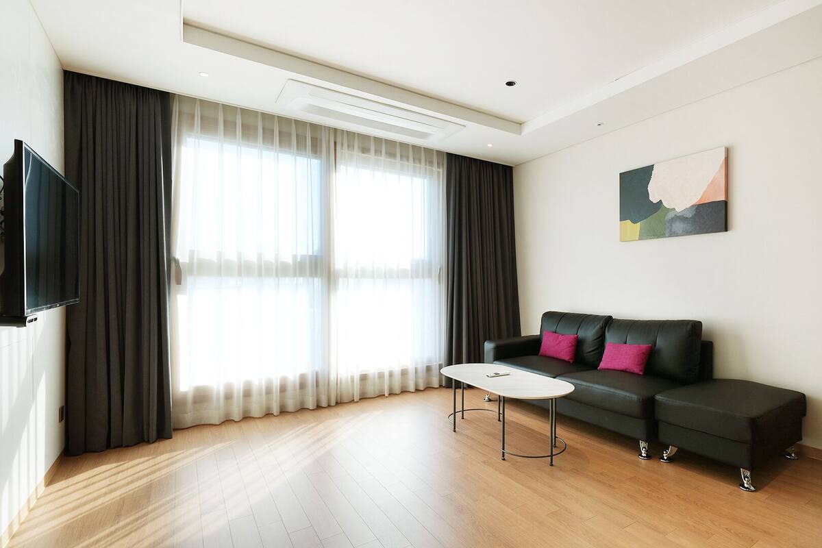 A modern living room with a black leather sofa, a white coffee table, and a large window with sheer curtains.