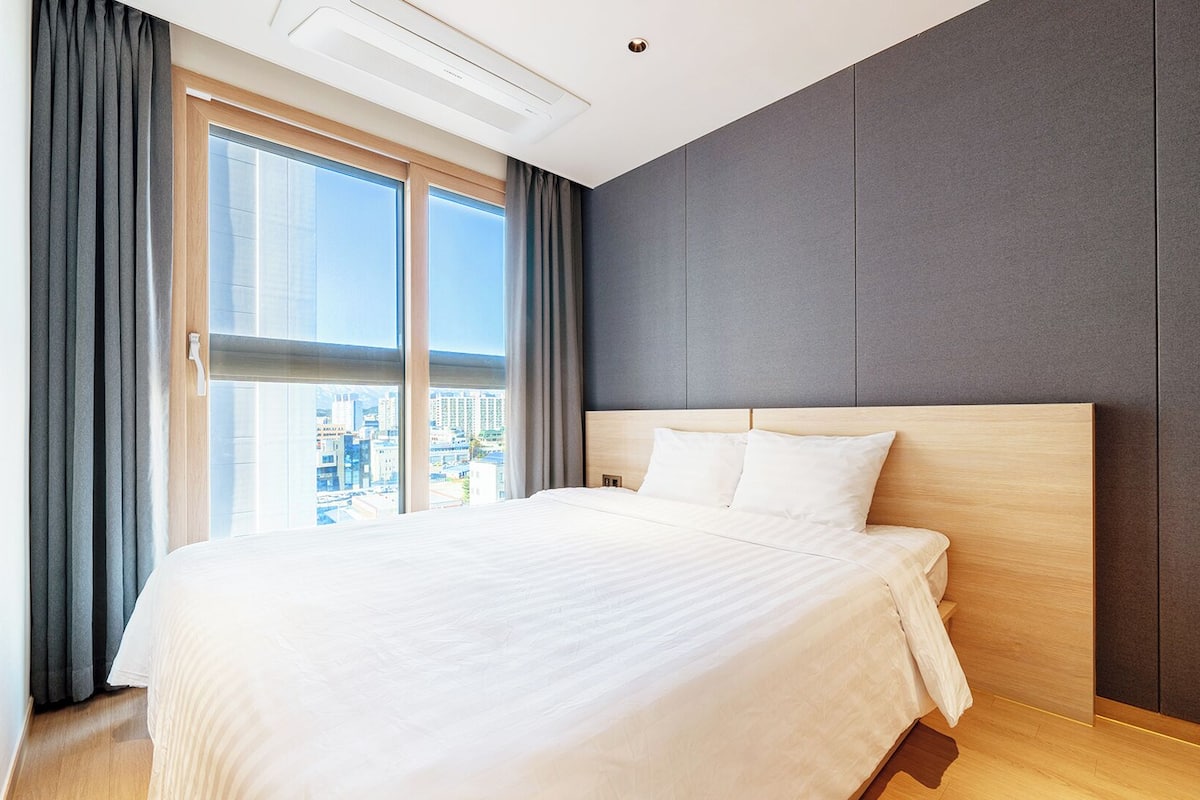 A modern hotel room with a large bed, a city view, and a grey accent wall.