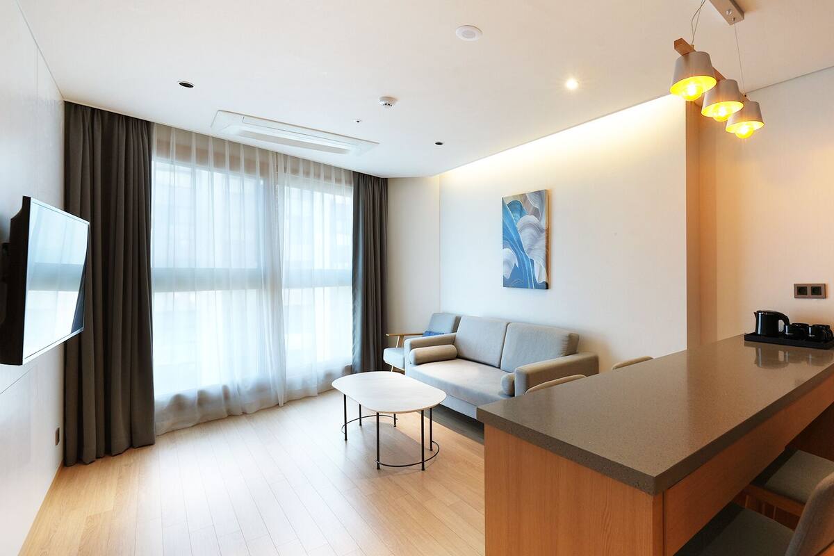 A modern hotel room with a flat-screen TV, a sofa, a dining area, and a painting on the wall.