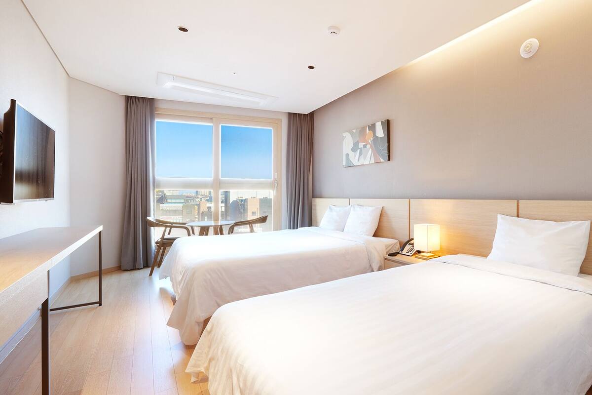 A hotel room with two beds, a desk, a chair, a TV, and a window with a city view.