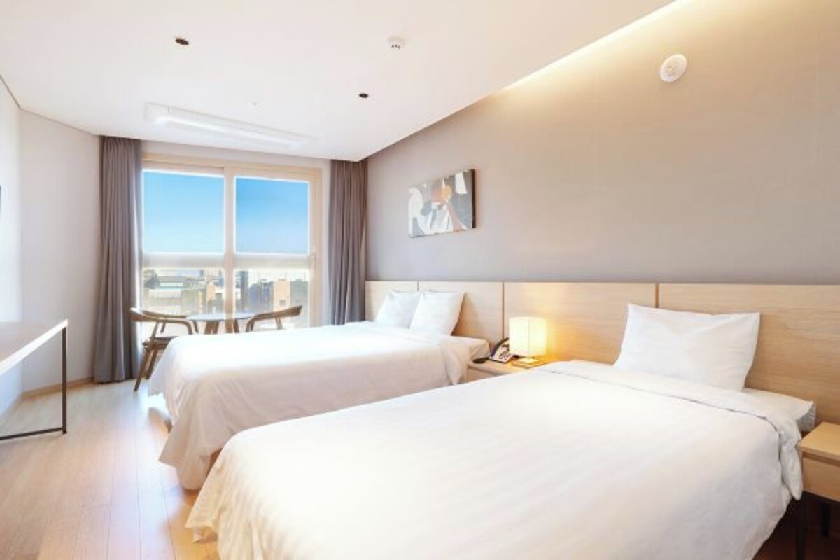A hotel room with two beds, a desk, and a window with a view of the city.