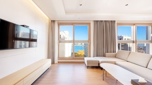A modern living room with a flat-screen TV mounted on the wall, a beige sofa, a small table, and large windows with curtains. - Dominiclava Sokco (Sokcho)