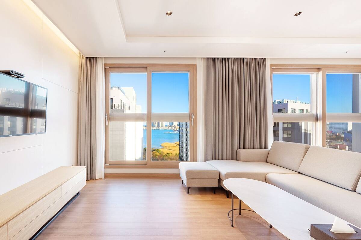 A modern living room with a flat-screen TV mounted on the wall, a beige sofa, a small table, and large windows with curtains.