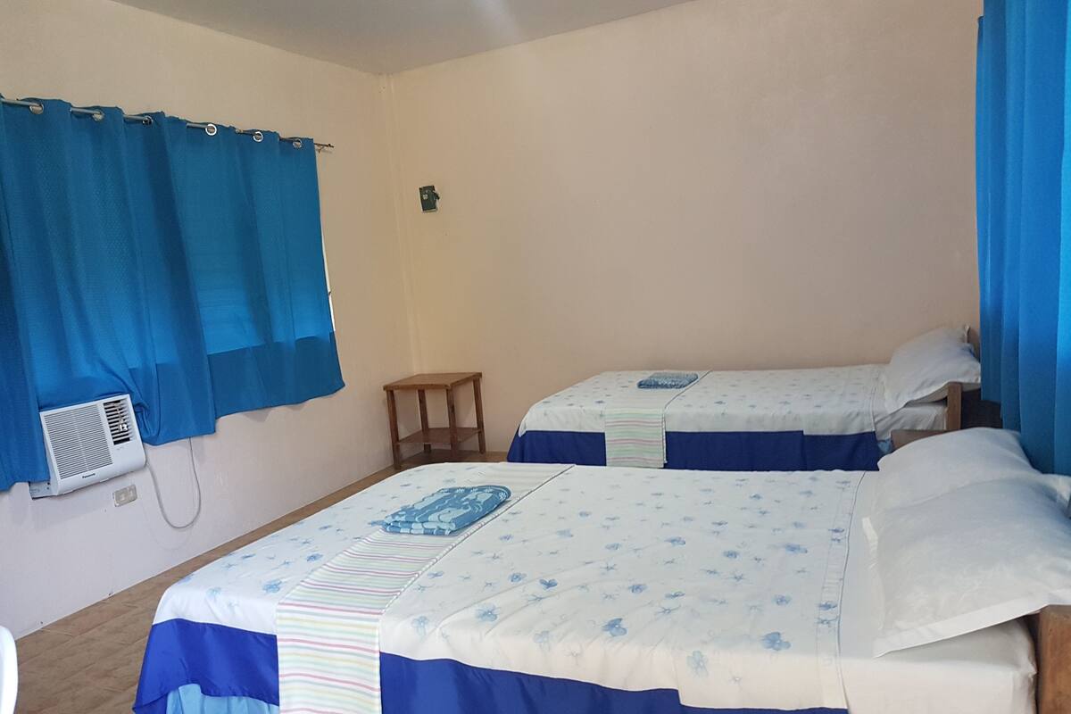 A room with two beds, each with a blue and white floral bedspread, a small wooden nightstand, and a wall-mounted air conditioning unit.