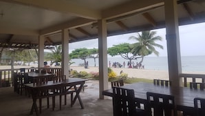 An outdoor seating area with wooden tables and chairs, overlooking a beach with palm trees and people.