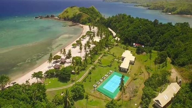 An aerial view of a coastal property with a beach, a tennis court, and lush greenery.
