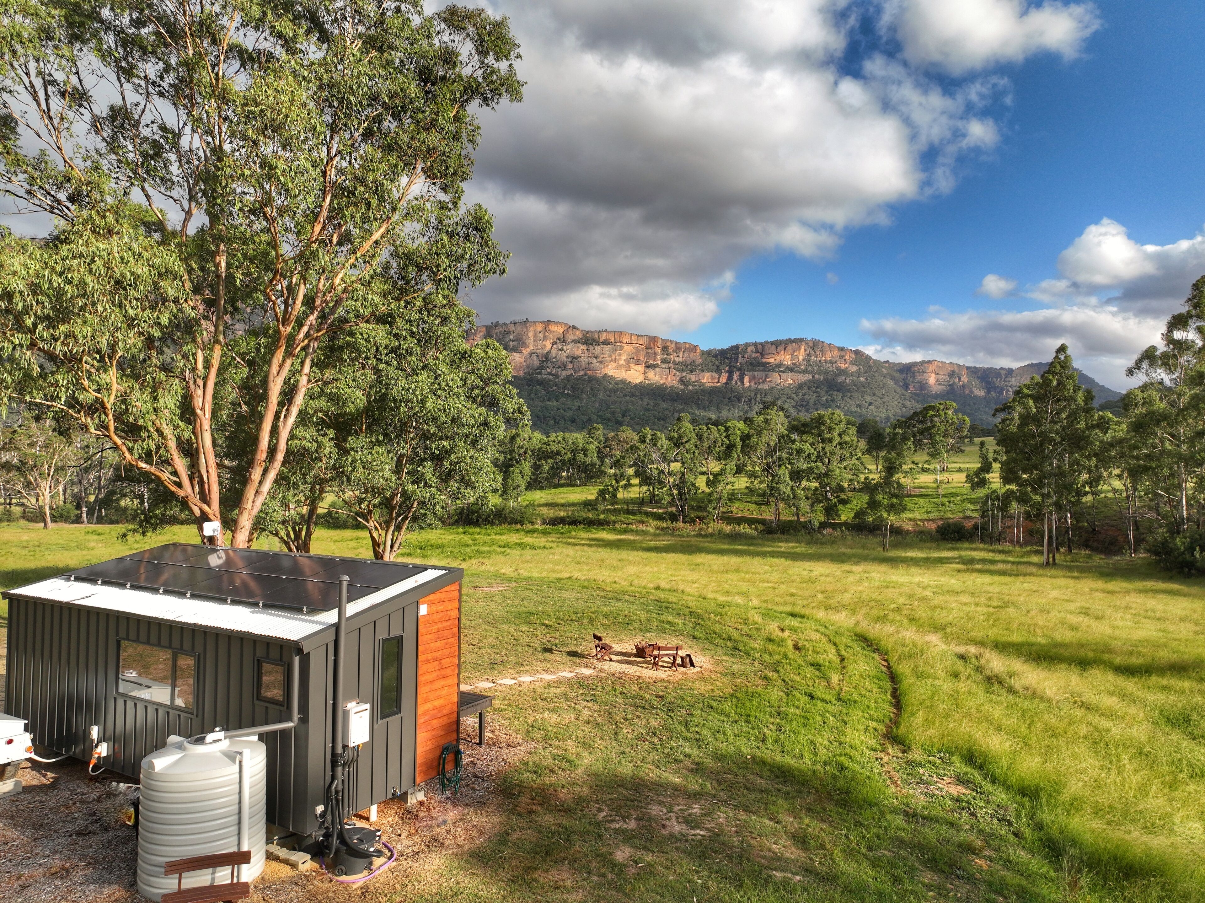 A small cabin with a metal roof and a wooden side, situated in a grassy field with trees and mountains in the background.
