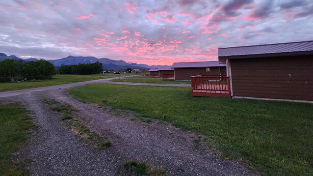 Entorno rural con un camino de grava que lleva a cabañas de madera, rodeado de vegetación y montañas al fondo.