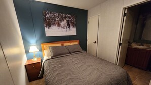 Un dormitorio con cama, mesita de noche con lámpara, armario y baño visible a través de una puerta abierta.