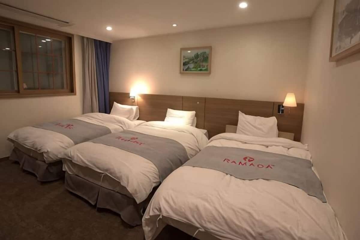 Habitación de hotel con tres camas, una ventana con cortinas, un cuadro en la pared y dos lámparas de pared.