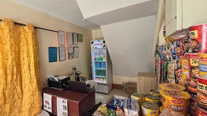 Ein kleiner Raum mit einem gelben Vorhang, einem mit Getränken gefüllten Kühlschrank, einem Automaten und verschiedenen Essensprodukten auf dem Boden.