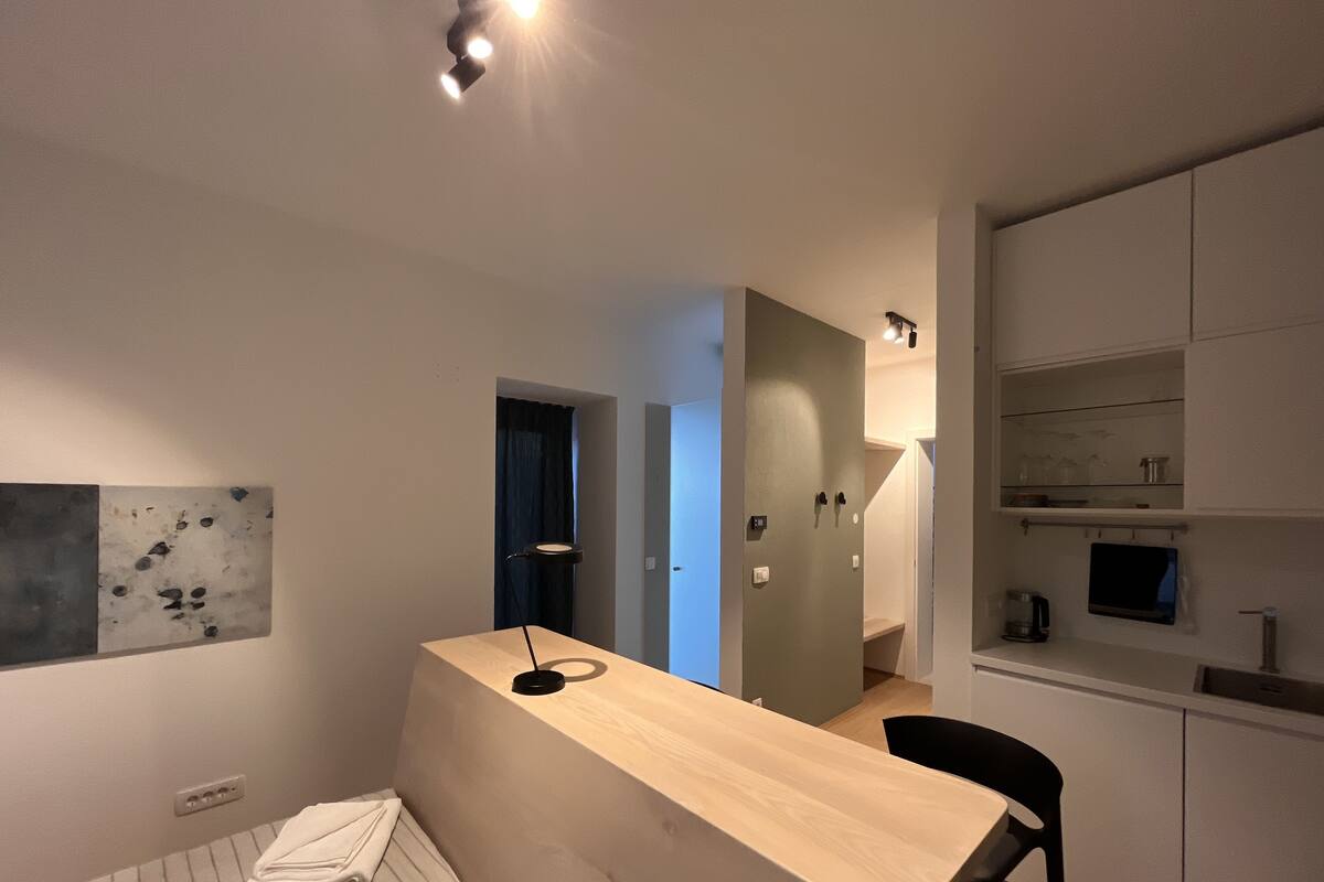 A modern, well-lit room with a wooden desk, a chair, a bed with a striped blanket, and a wall-mounted artwork.