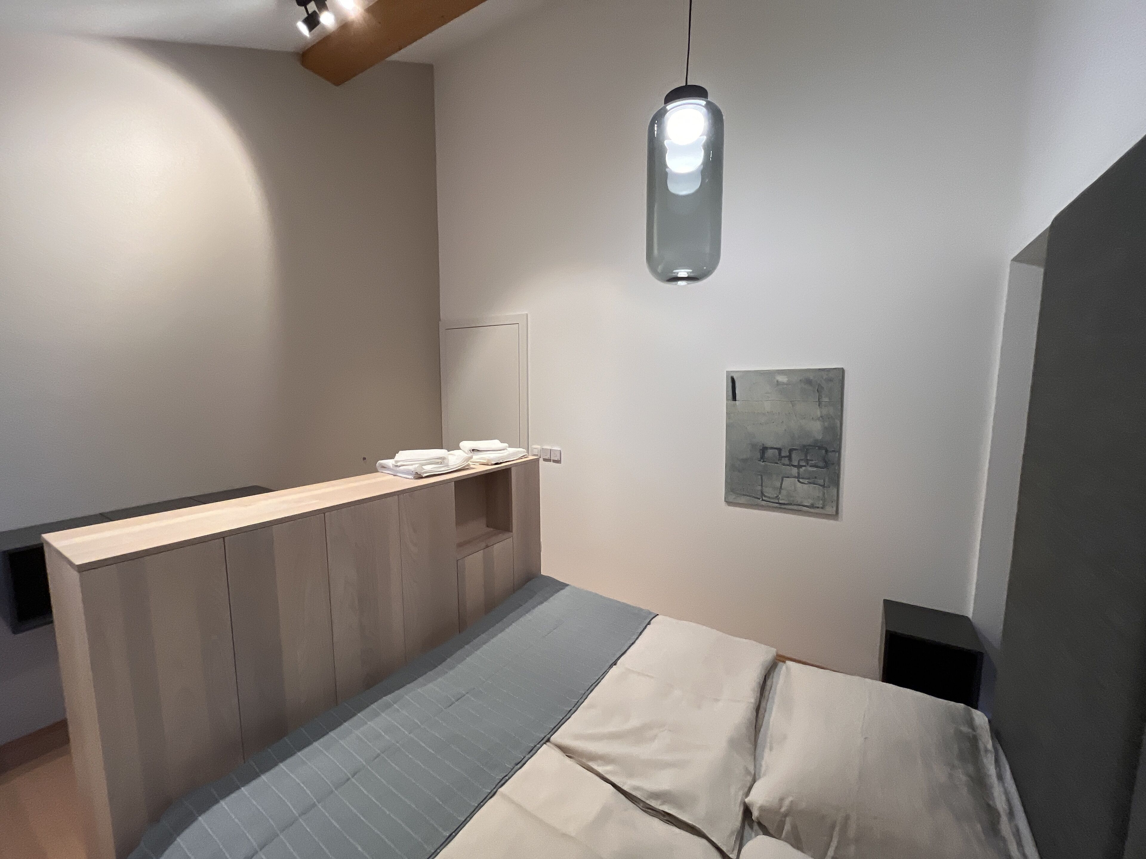 Un dormitorio moderno con una cama de madera, una mesita de noche con toallas, una lámpara colgante y una obra de arte en la pared.
