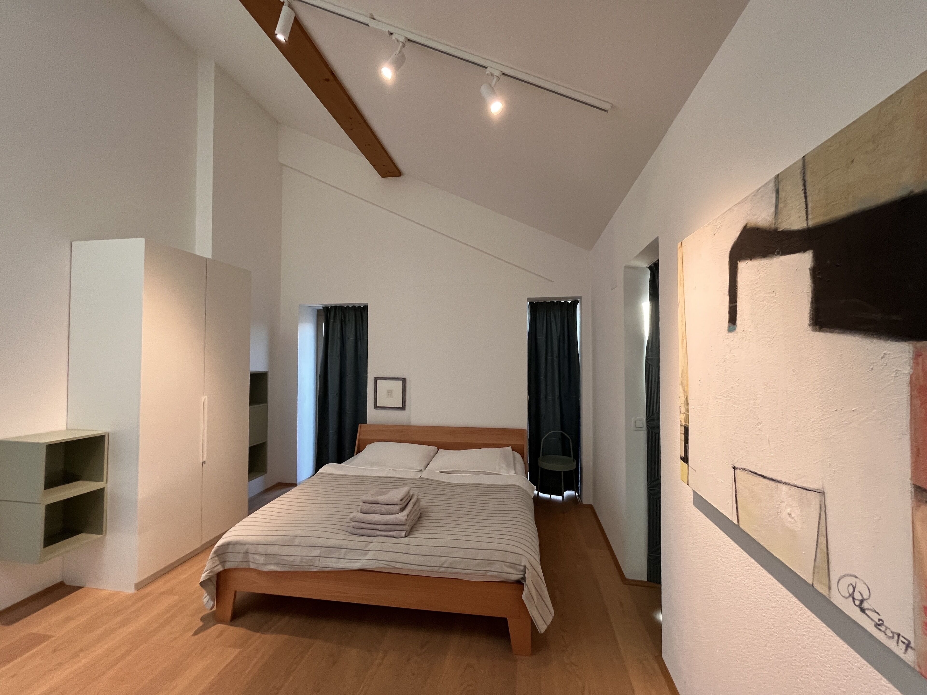 A modern bedroom with a wooden bed, a wardrobe, a small seating area, and a painting on the wall.
