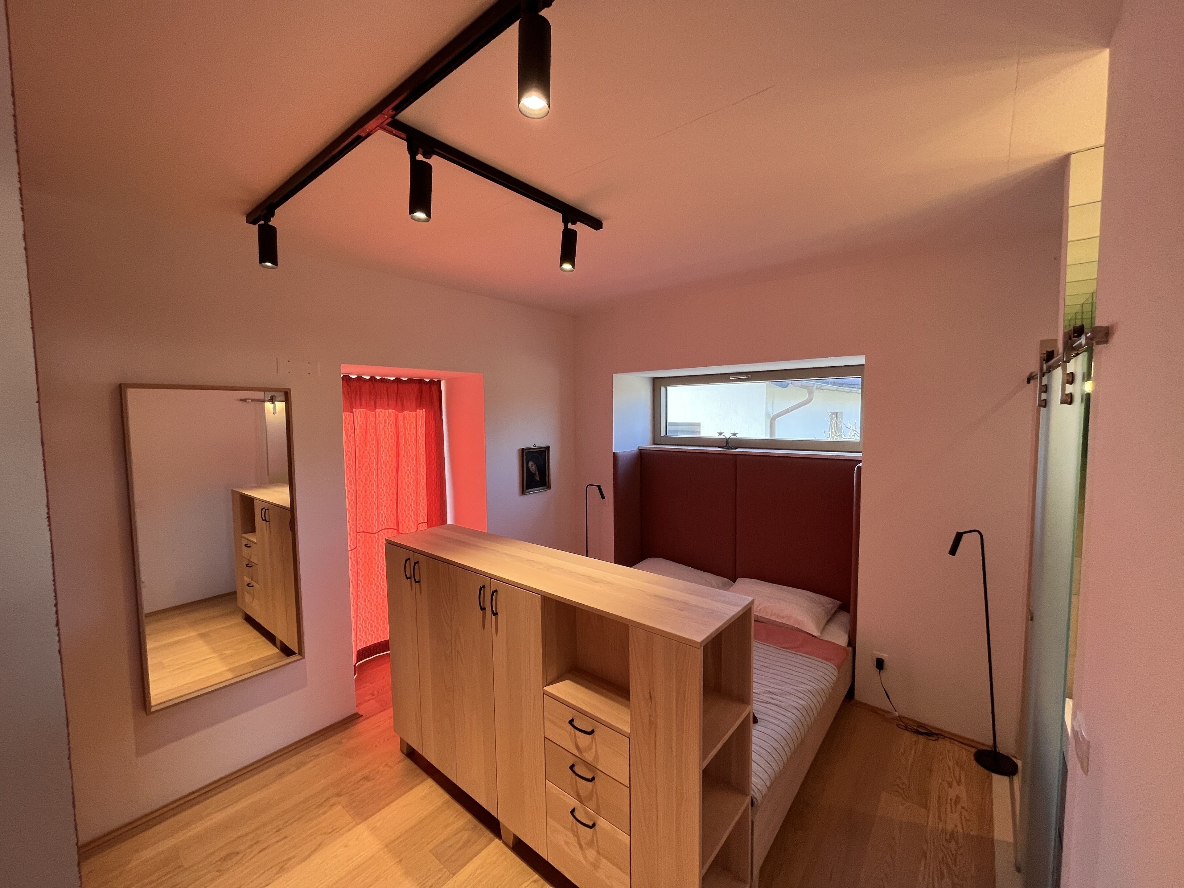 Un dormitorio con cama, armario, espejo, ventana y lámpara en el techo.