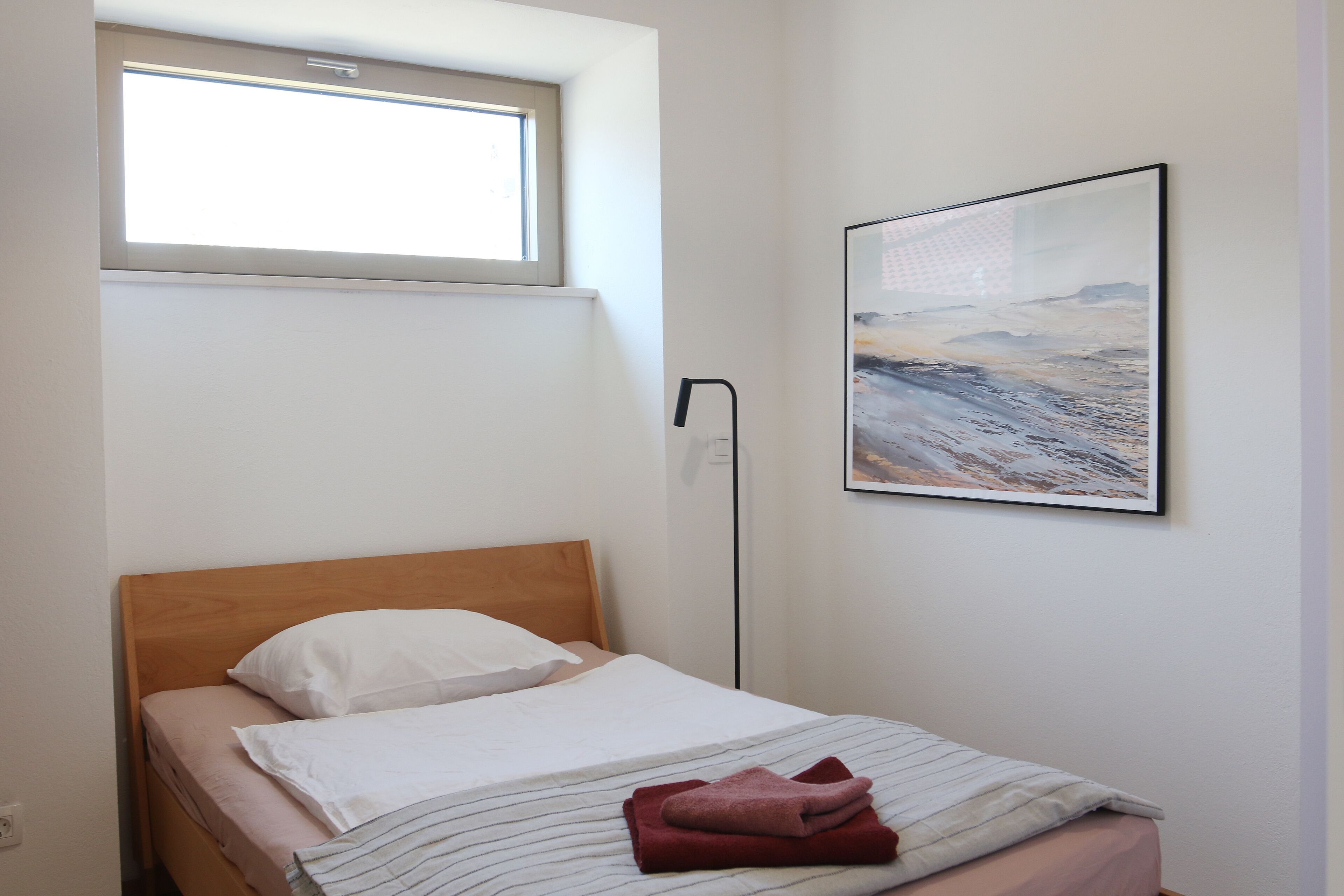 A single bed with a wooden headboard, white bedding, and a red towel on the footboard. A window above the bed allows natural light, and a framed abstract painting hangs on the wall.