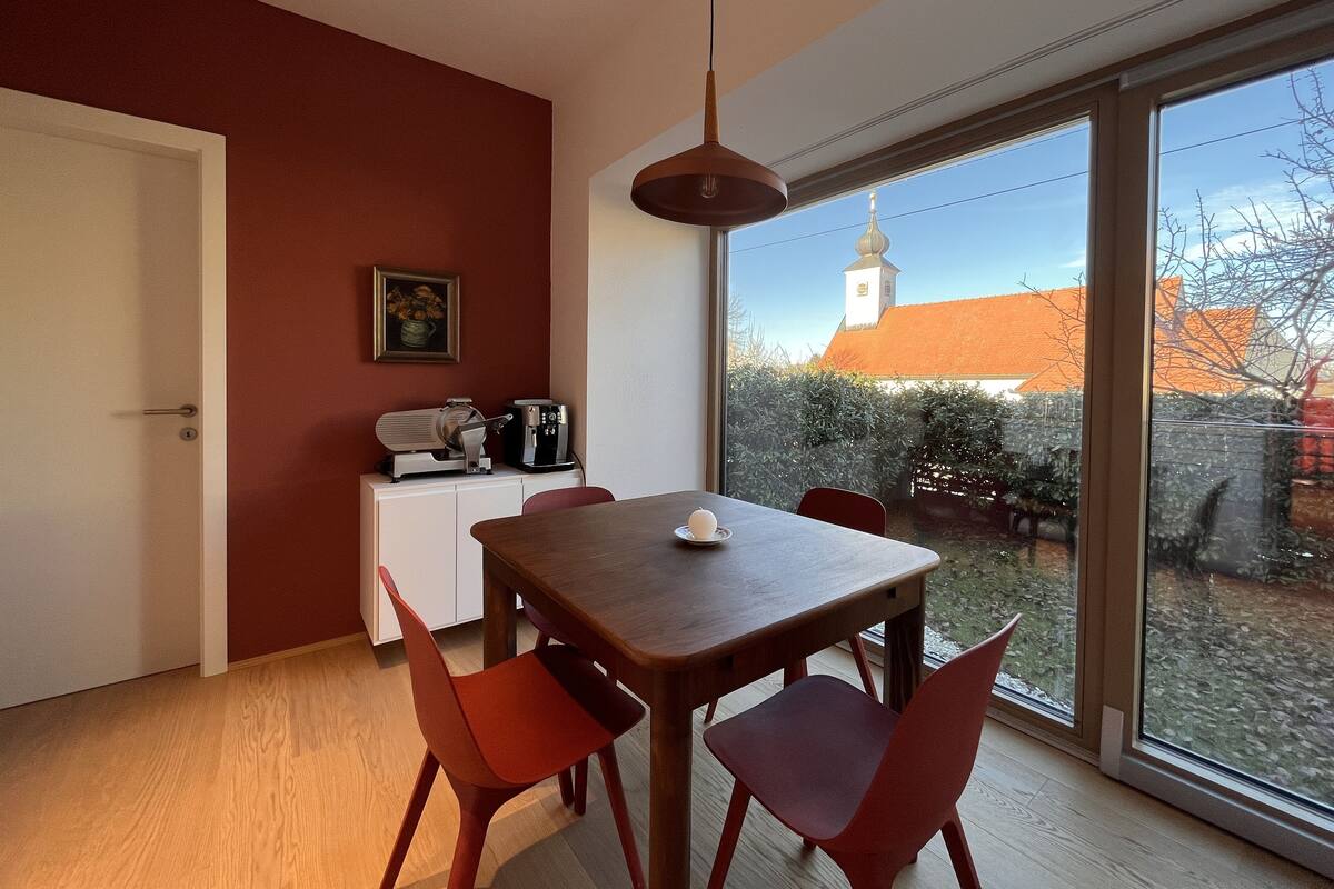 A dining area with a wooden table and red chairs, a white cabinet with kitchen appliances, and a large window offering a view of a building with a tower.